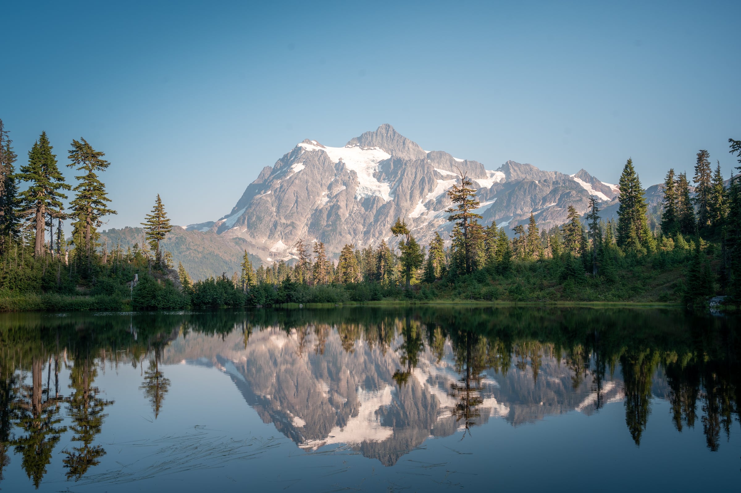 A rugged snow-capped mountain and surrounding forest reflect in a still alpine lake.