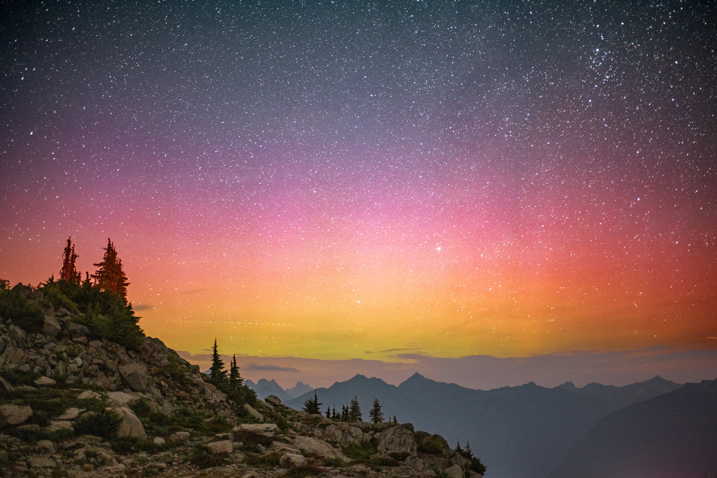 Bands of pink, orange, and green aurora glow above a rocky mountain horizon under dense stars.