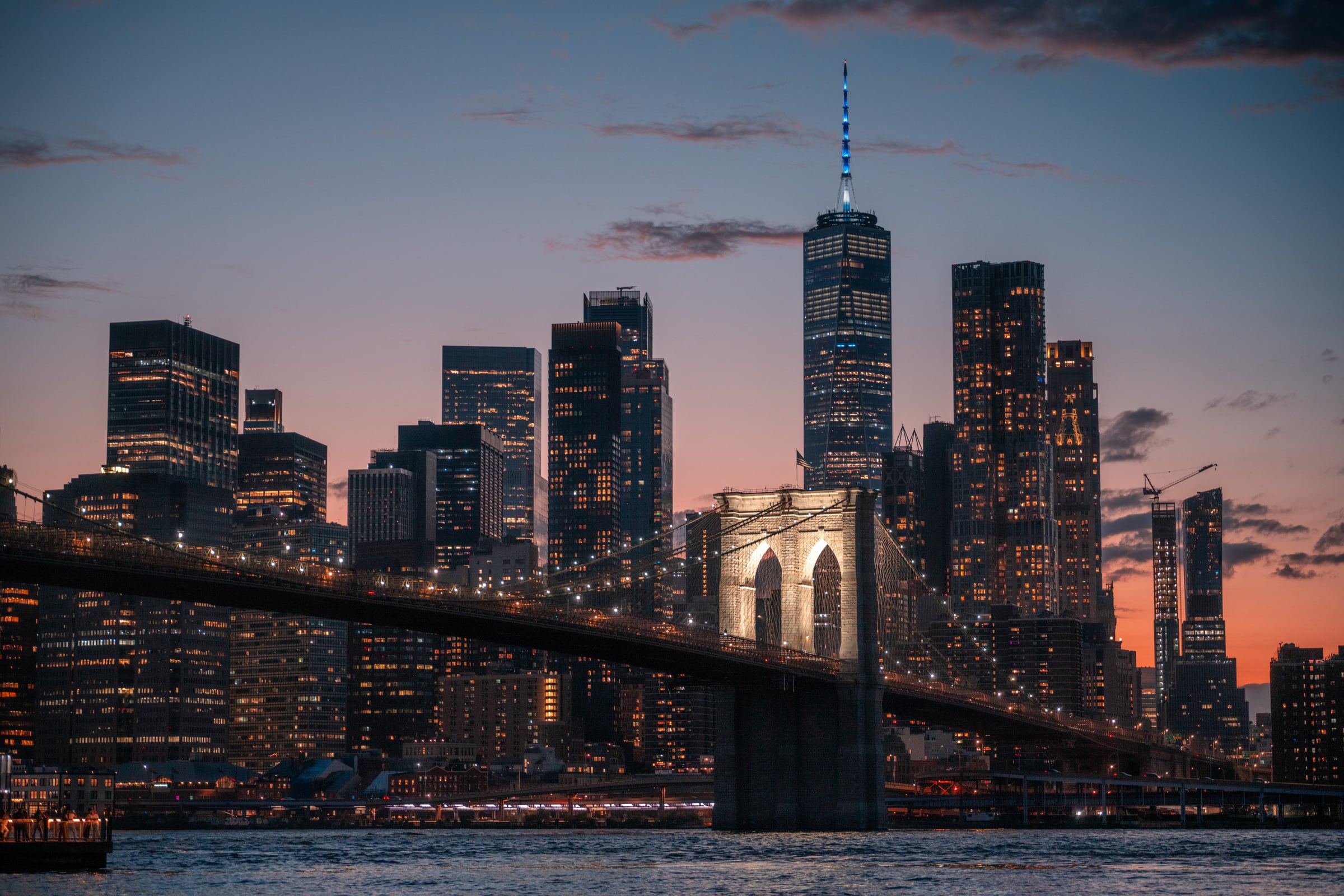 The Brooklyn Bridge crosses a dusk skyline centered on One World Trade Center.