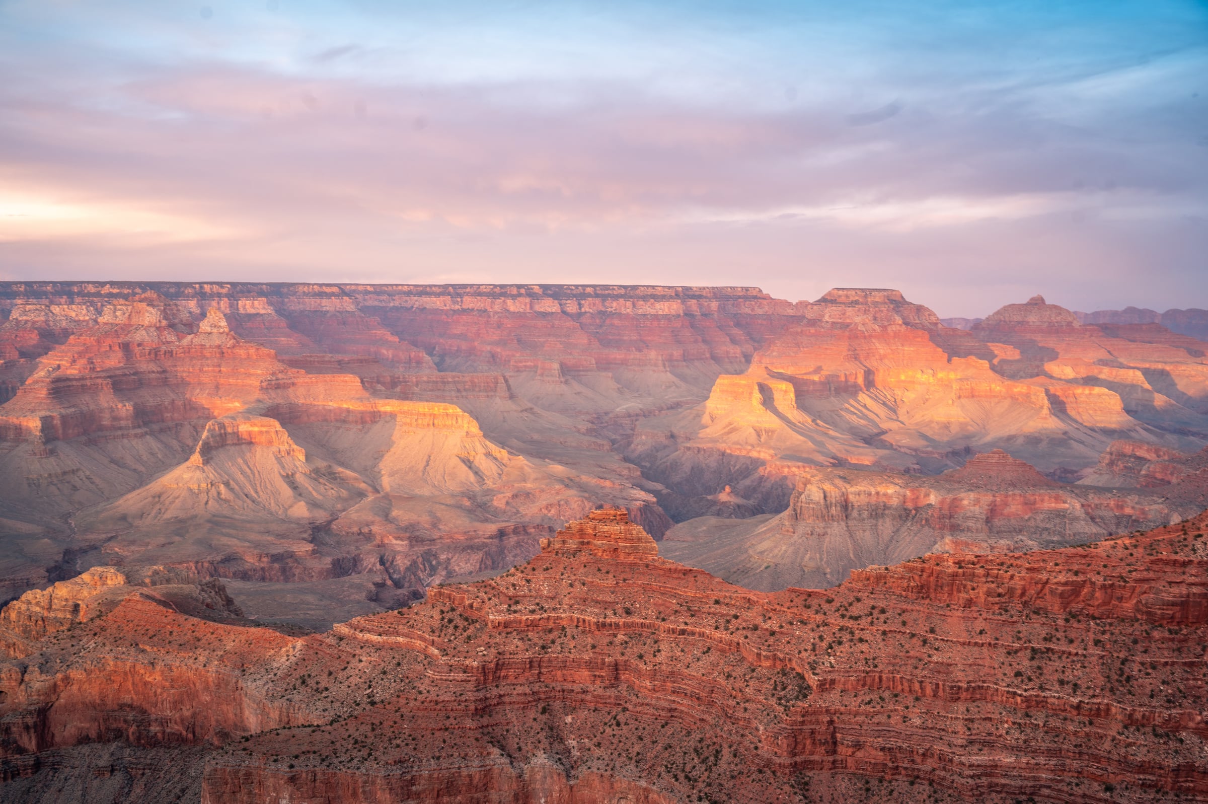 Sunset light washes layered red cliffs and mesas beneath a pastel sky.
