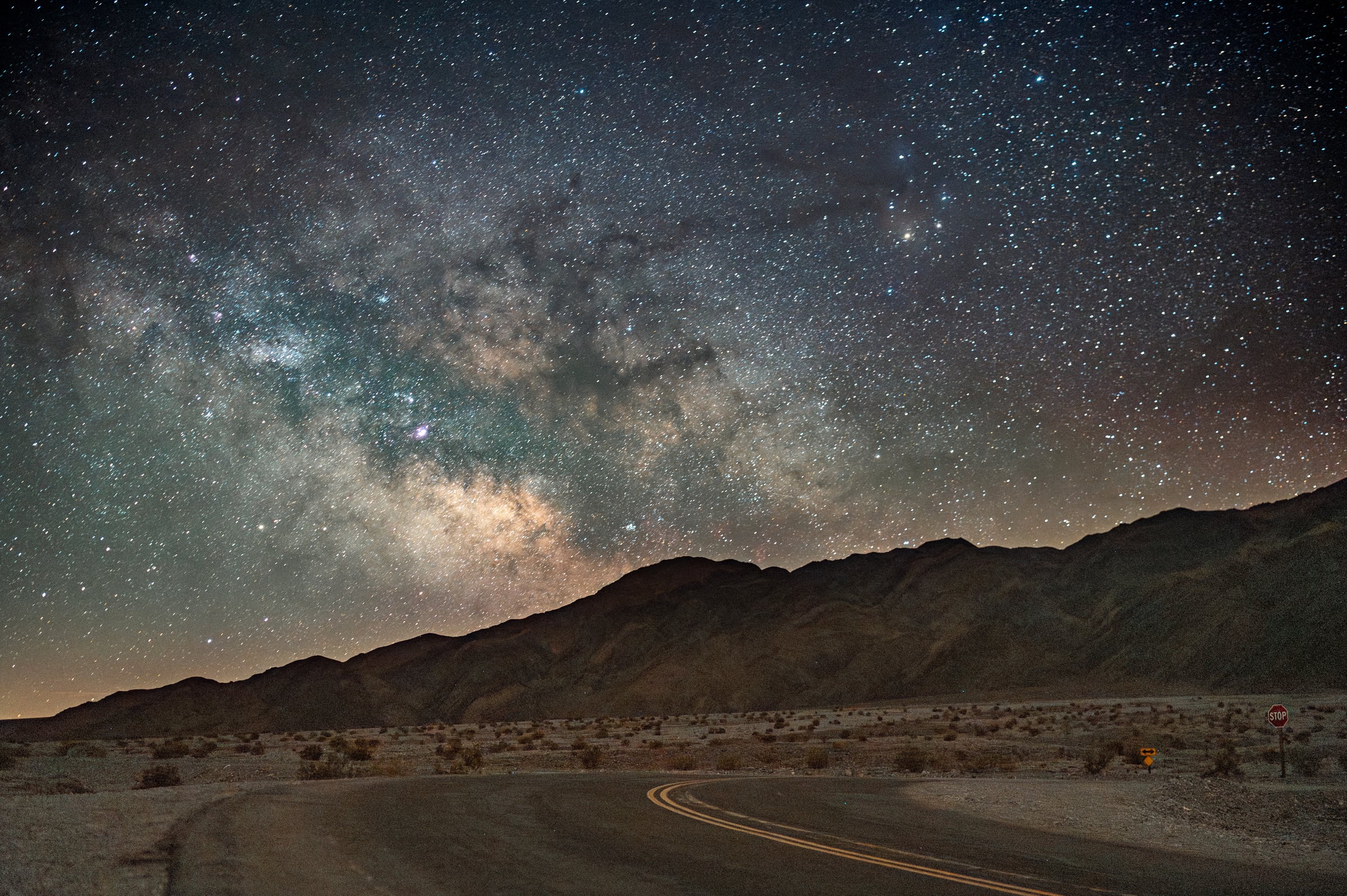 The Milky Way arcs over a winding desert road and dark mountain ridge.