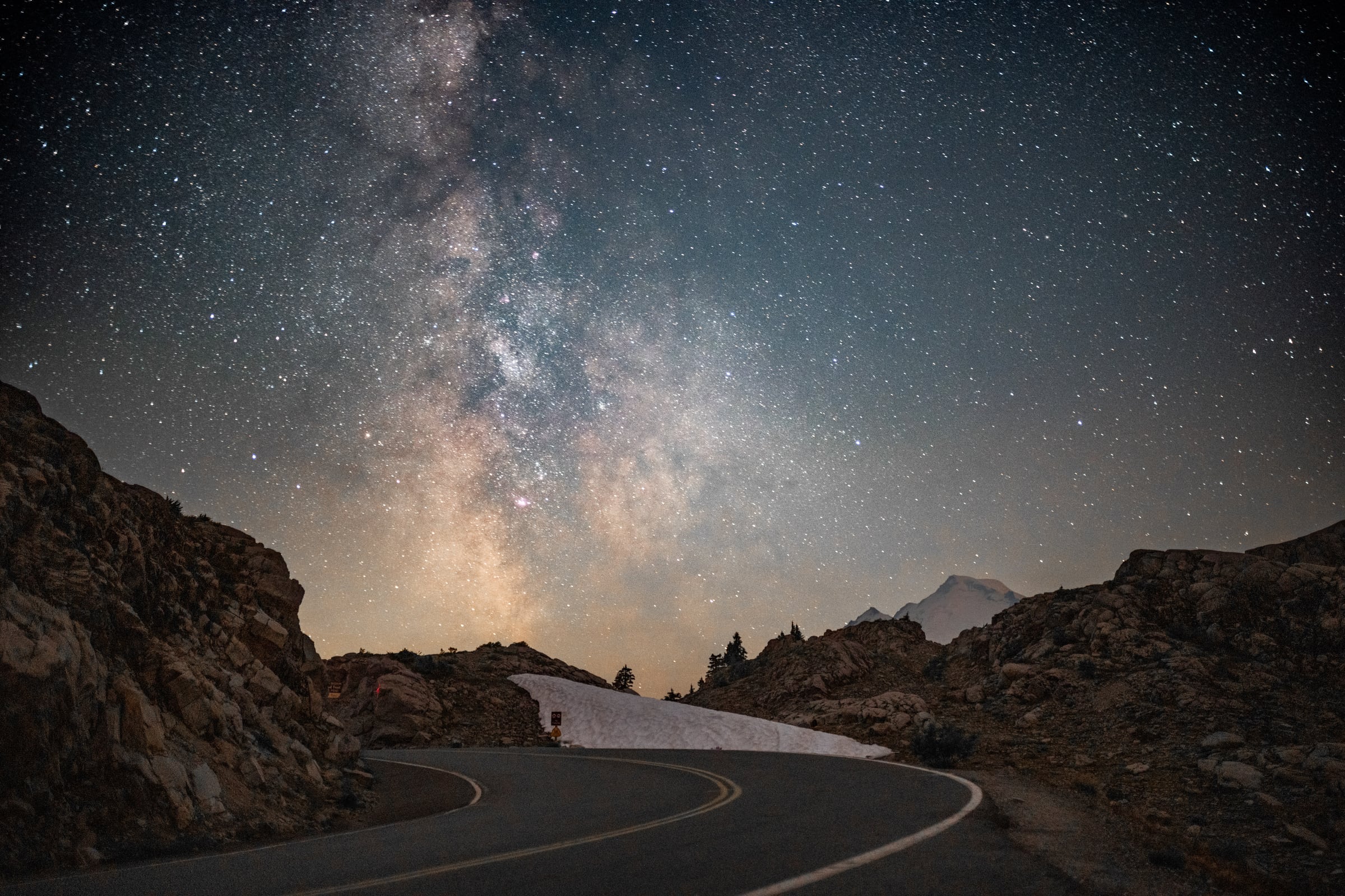 The Milky Way rises above a curving mountain road bordered by snow and rock.