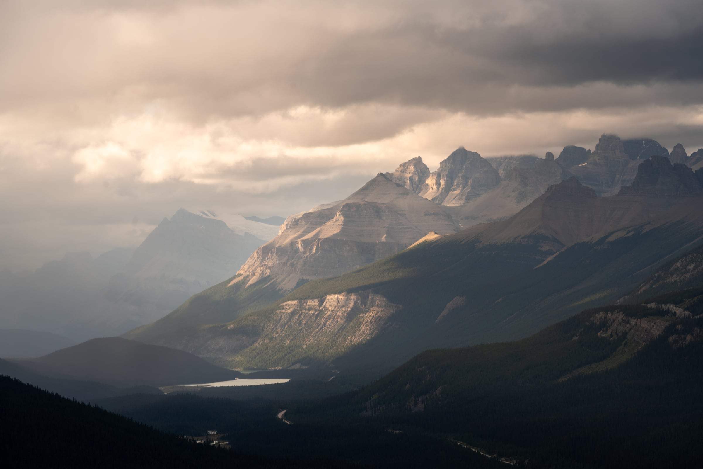 A shaft of late light reaches jagged mountain peaks above a shadowed valley and lake.