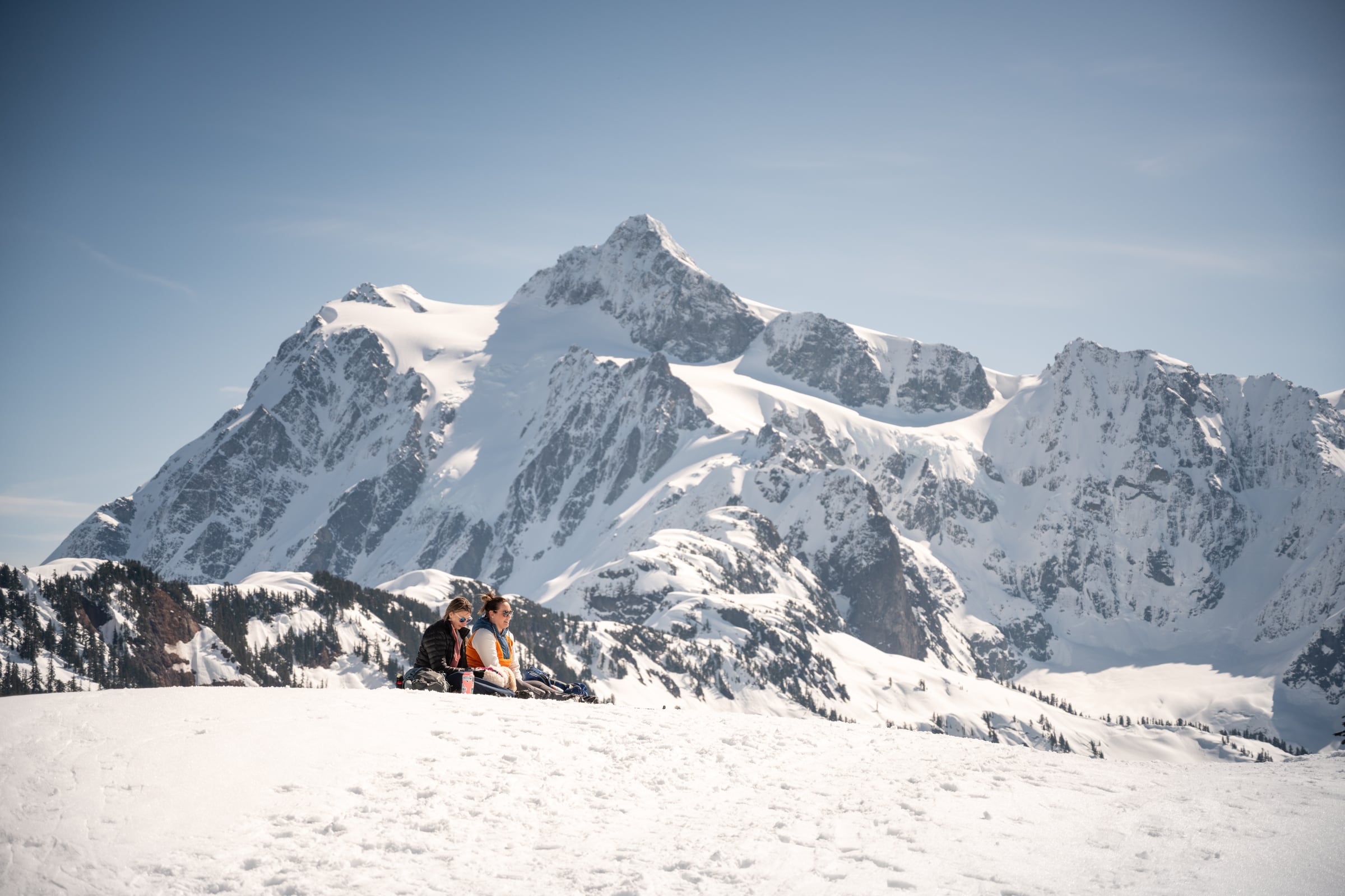 Two people sit on a bright snowfield beneath a vast wall of jagged white peaks.