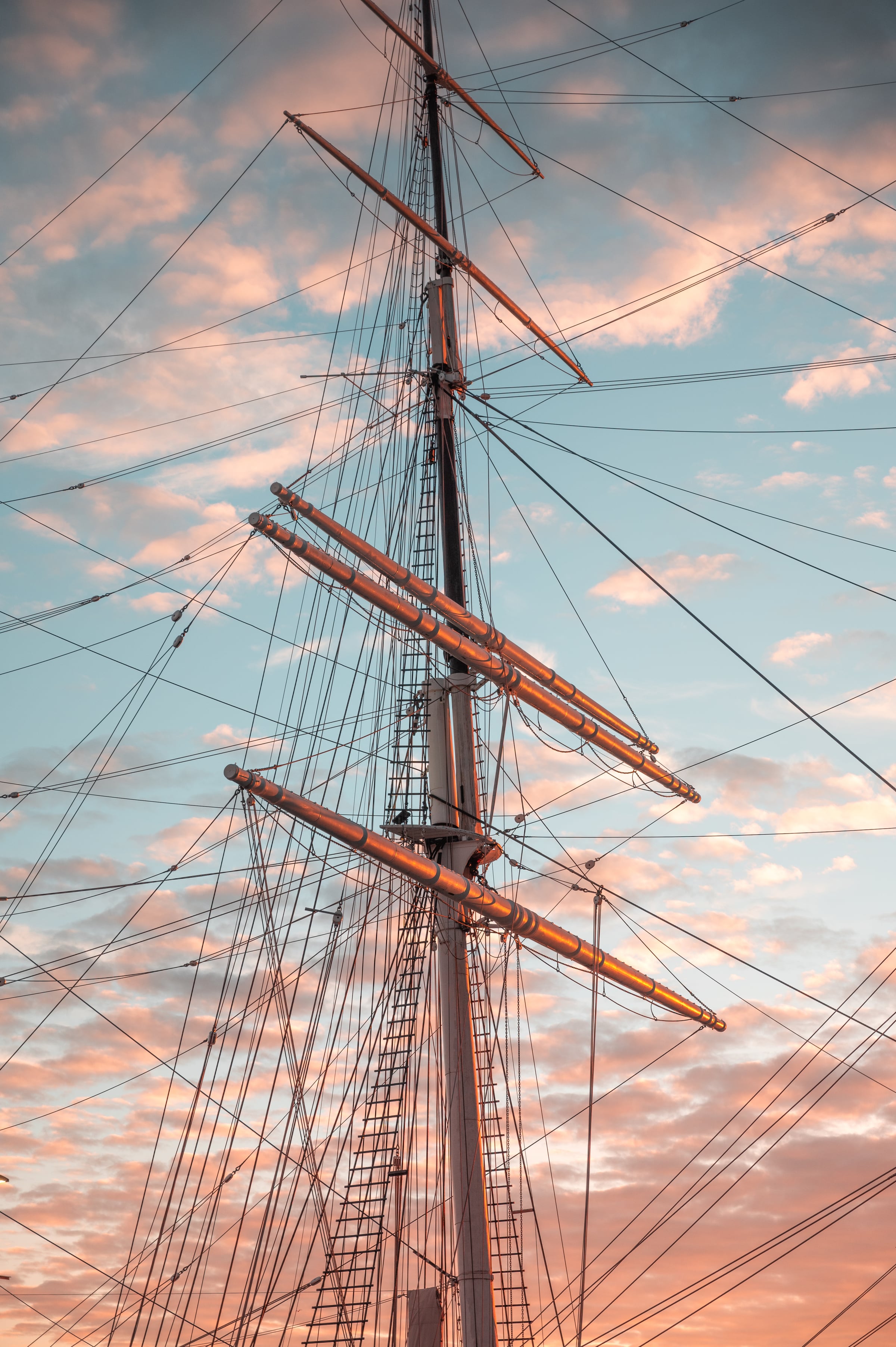 Tall-ship masts and rigging form a lattice against peach and blue evening clouds.
