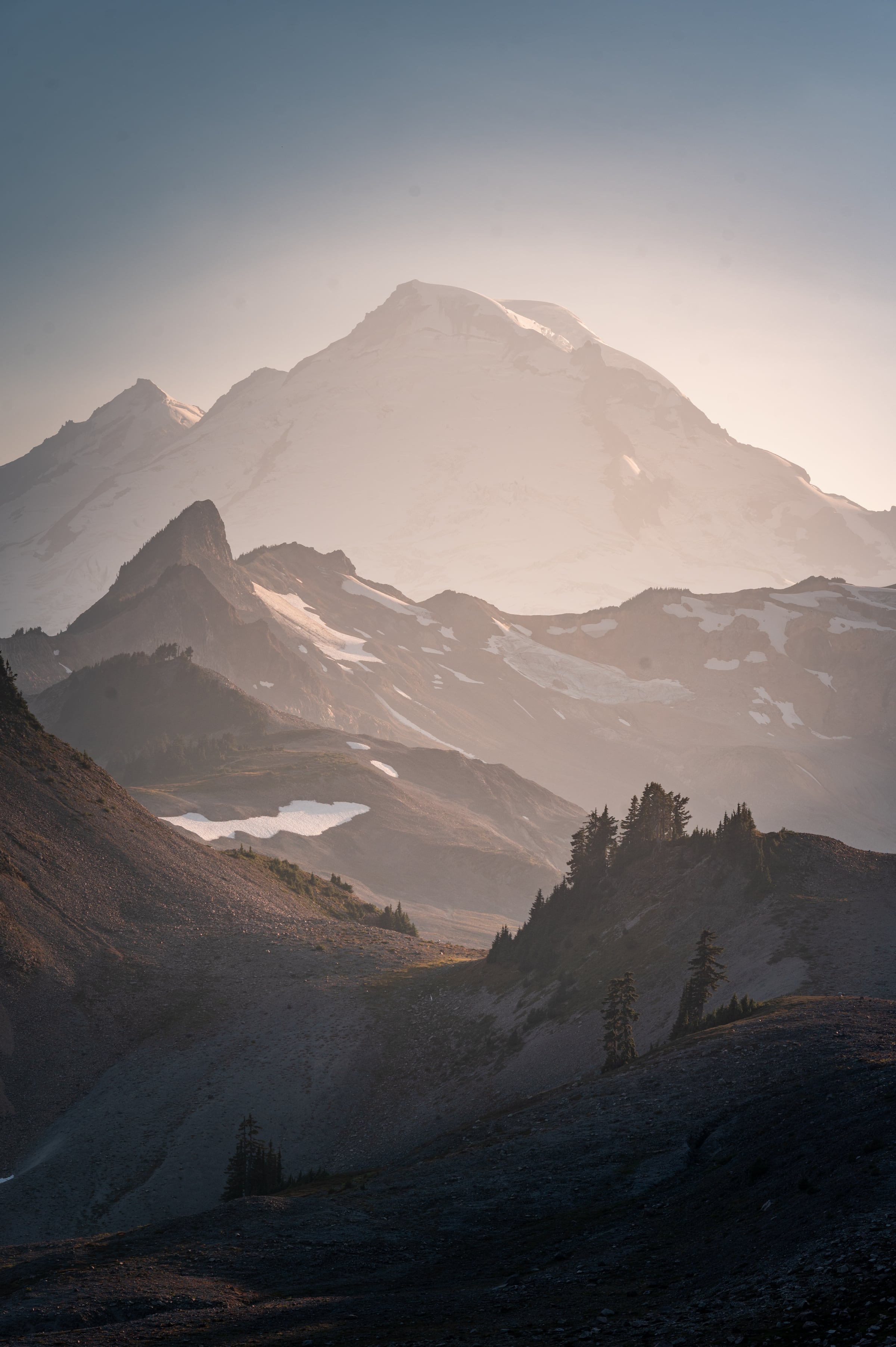 A snow-covered volcanic peak rises through warm haze beyond dark rocky ridges.