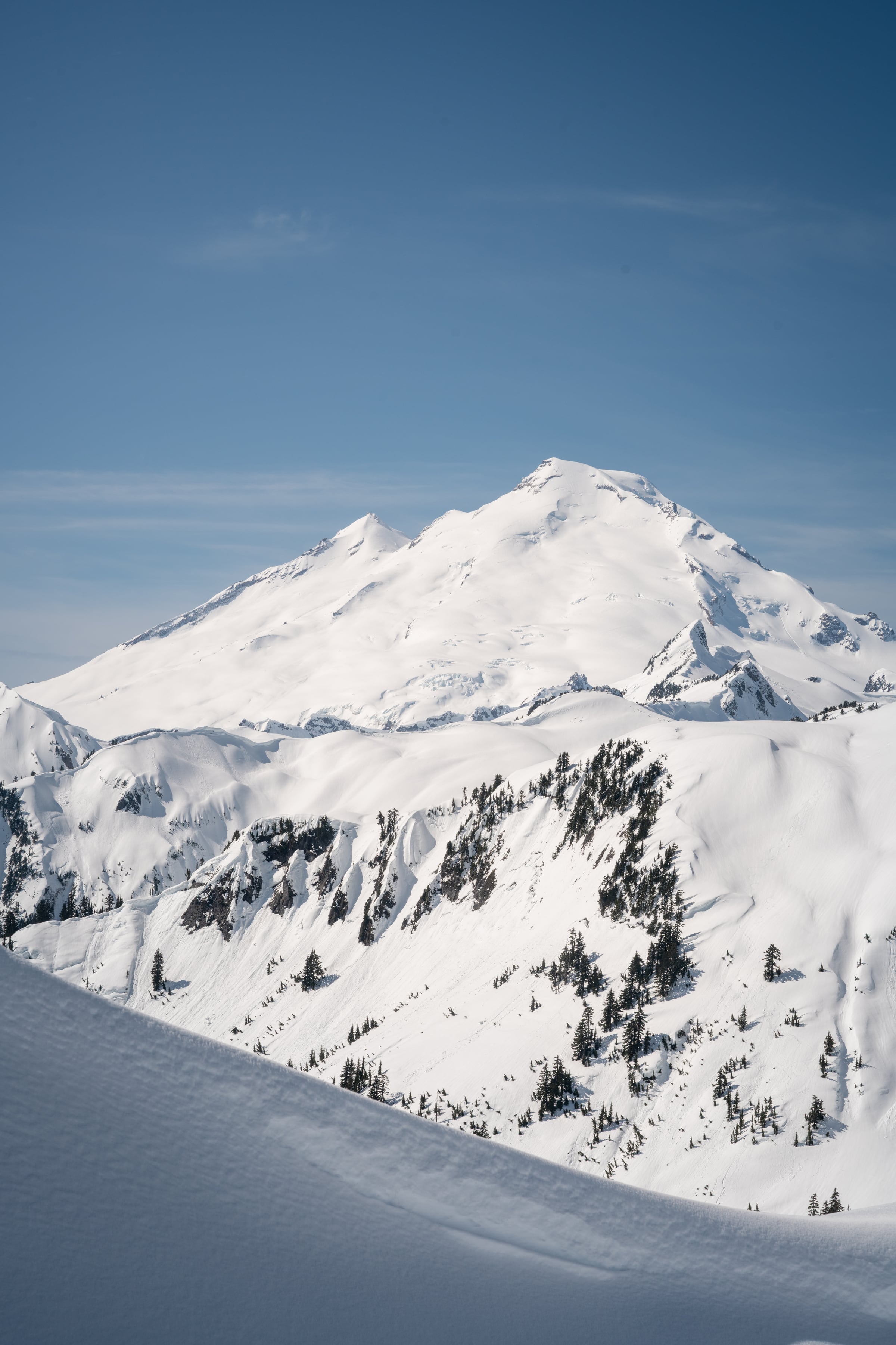 A solitary snow-covered volcanic peak rises above rolling white ridges beneath a deep blue sky.